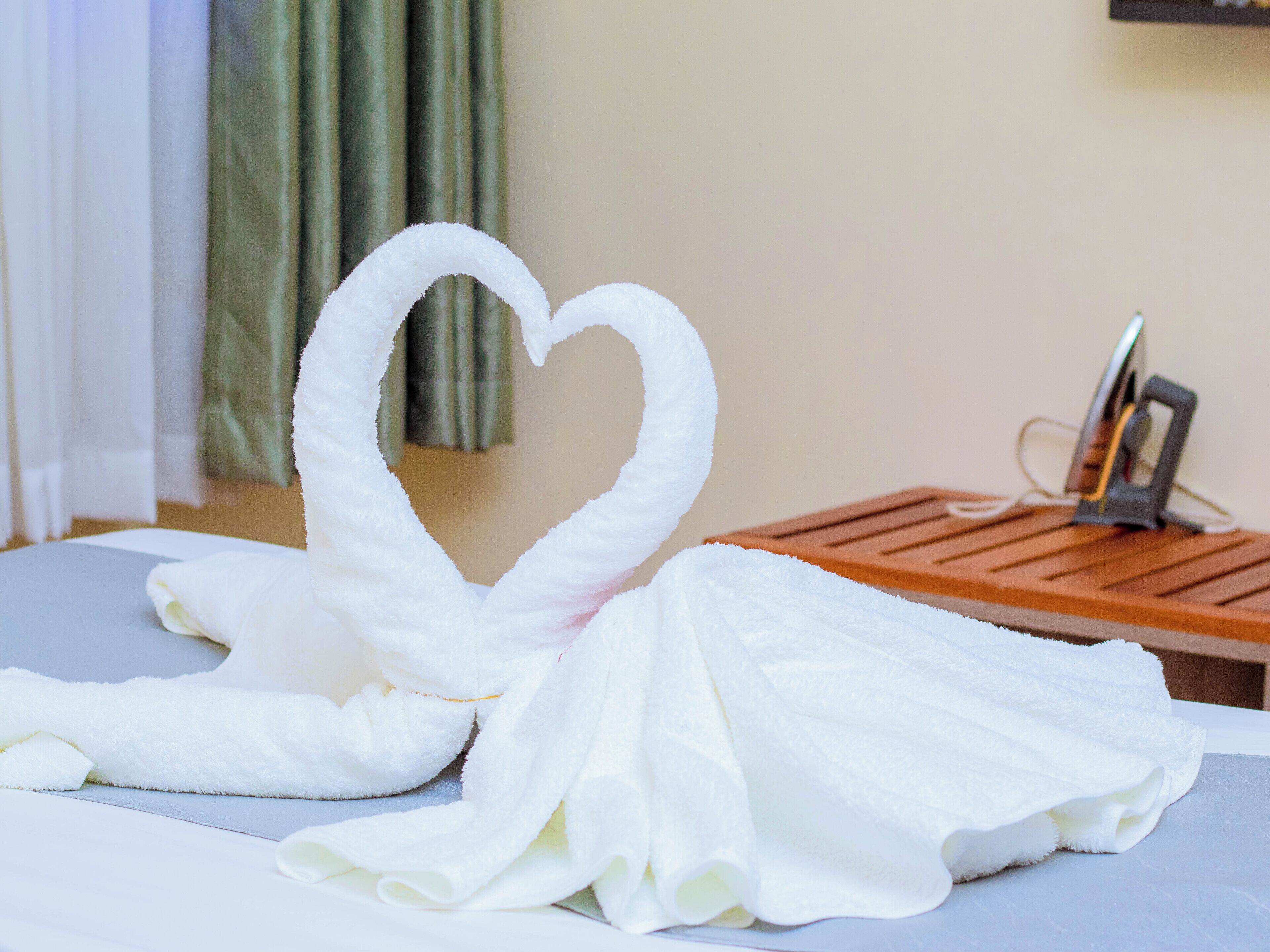 Uma cama com arranjo de toalha em formato de coração e uma mesa de cabeceira com carregador de celular.