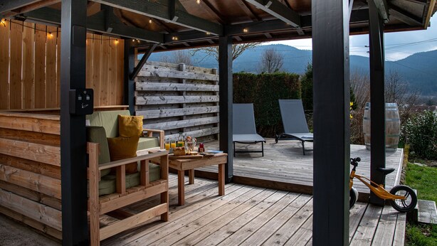 A wooden deck with a sofa and chairs, a table with drinks and snacks, and a bicycle. - Gîte chez Alice et Marcel: sauna, cabin, chicken coop, bicycles & garden. (Saulxures-sur-Moselotte)