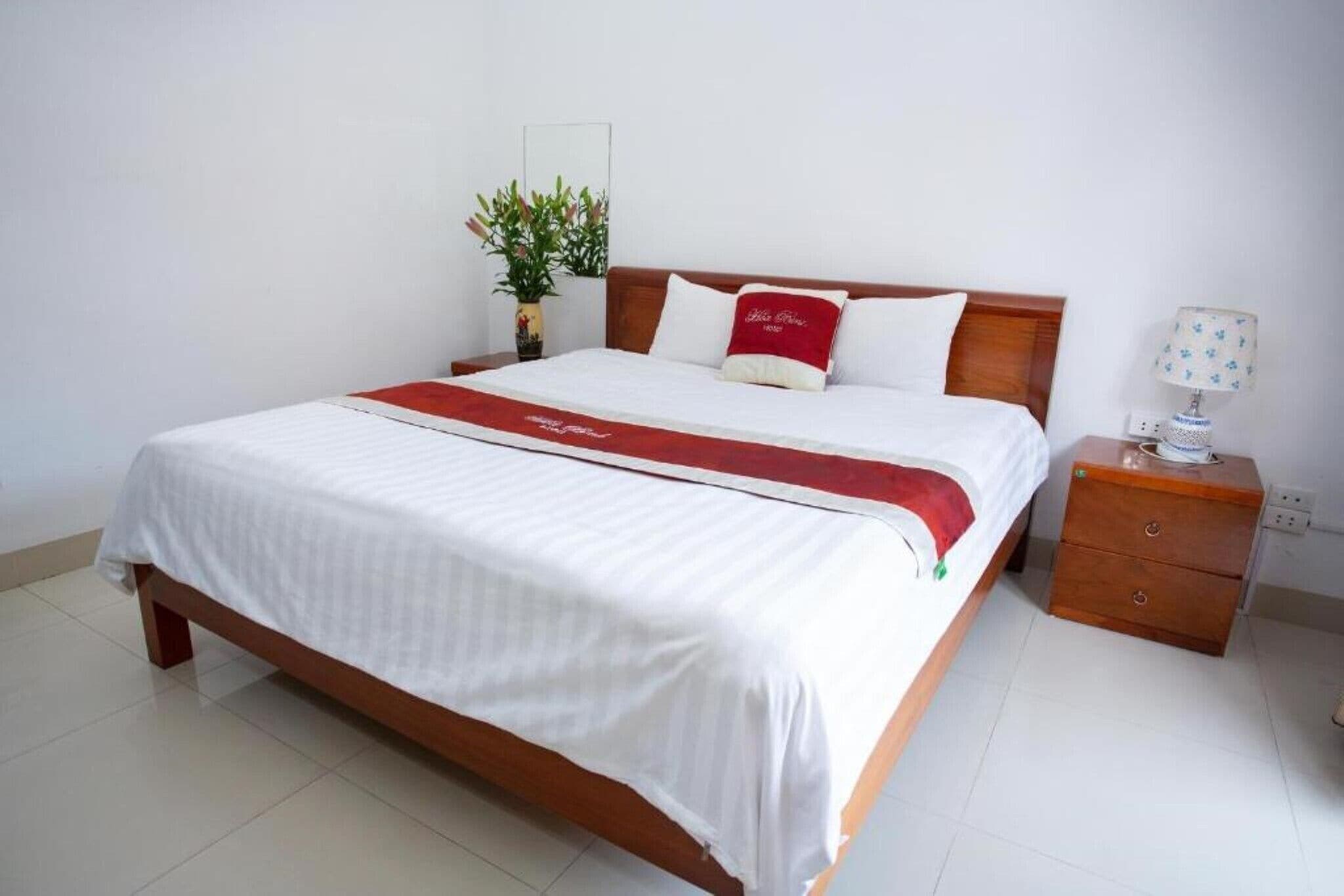 A hotel room with a wooden bed, white bedding, a red throw, a bedside table with a lamp, and a small plant.