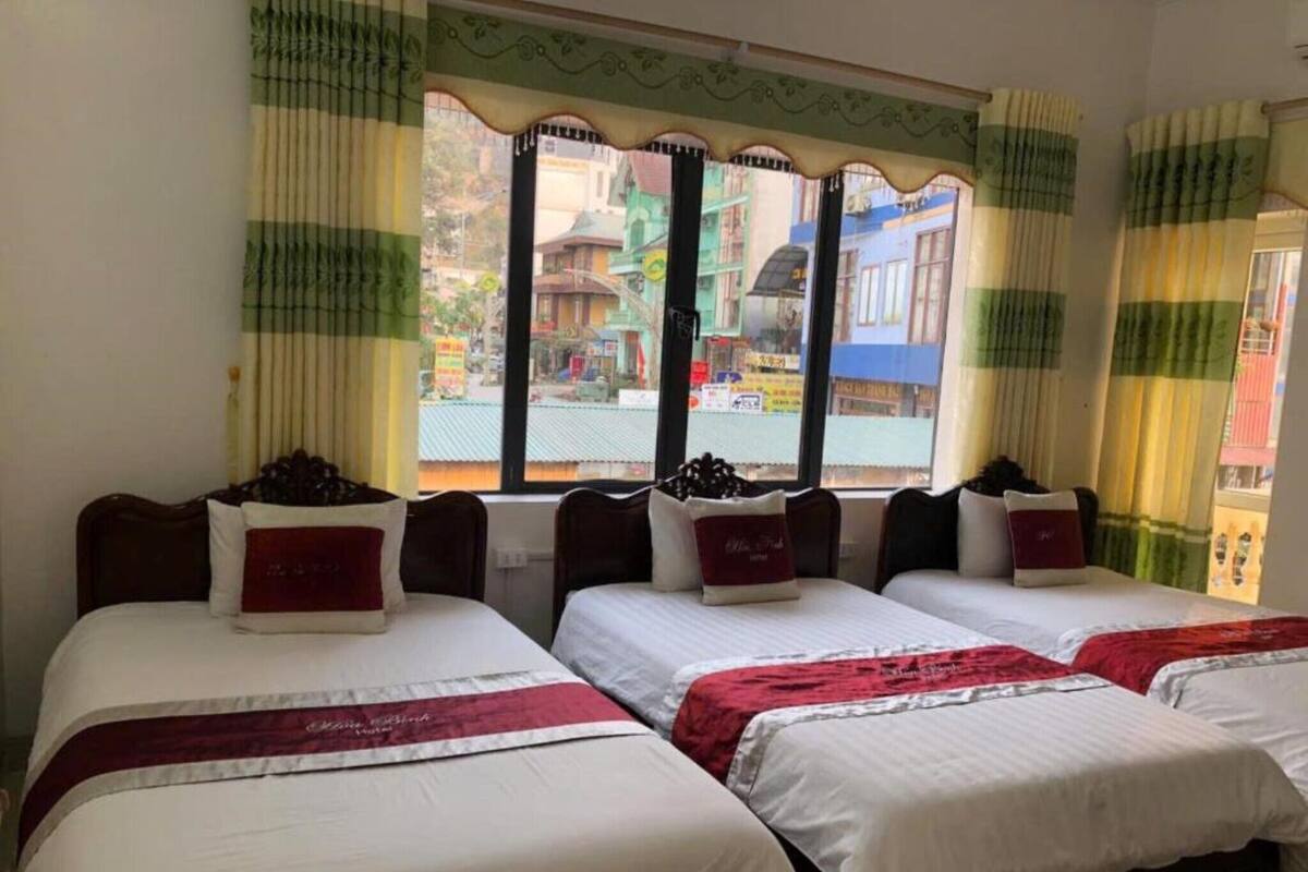 A hotel room with three beds, each with a white bedspread and a red accent pillow. The room has a large window with green and yellow striped curtains, and a view of buildings outside.