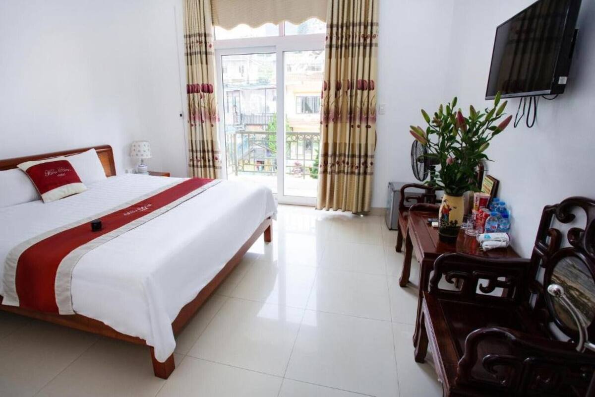 A hotel room with a large bed, a desk with a chair, a television mounted on the wall, and a balcony with curtains.