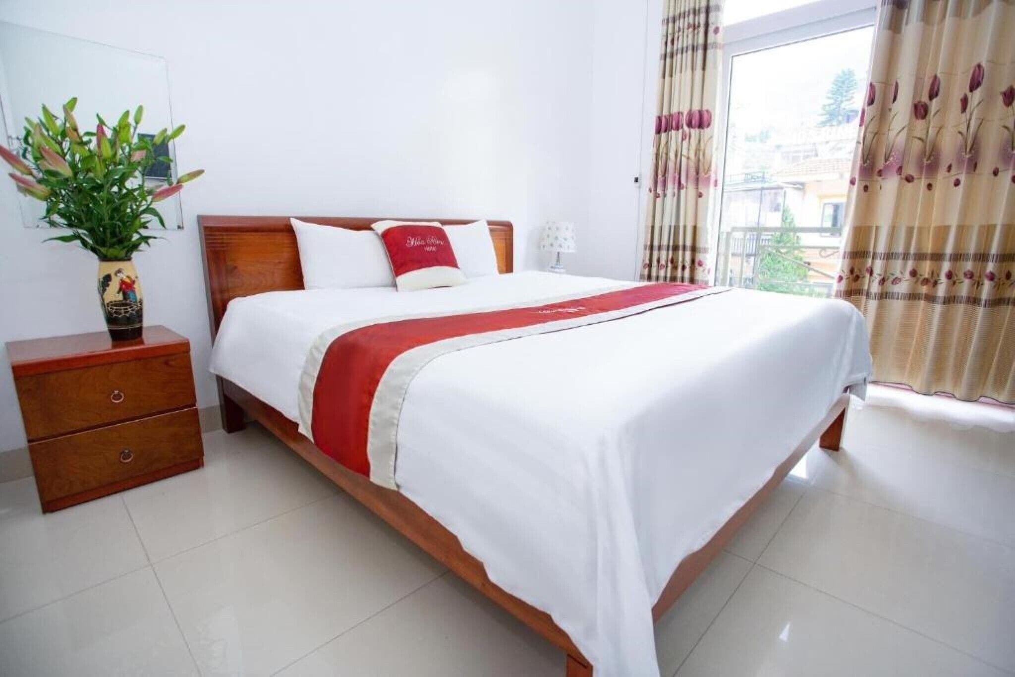 A neatly arranged bedroom with a wooden bed, white bedding, a red throw, a bedside table with a vase, and a window with curtains.