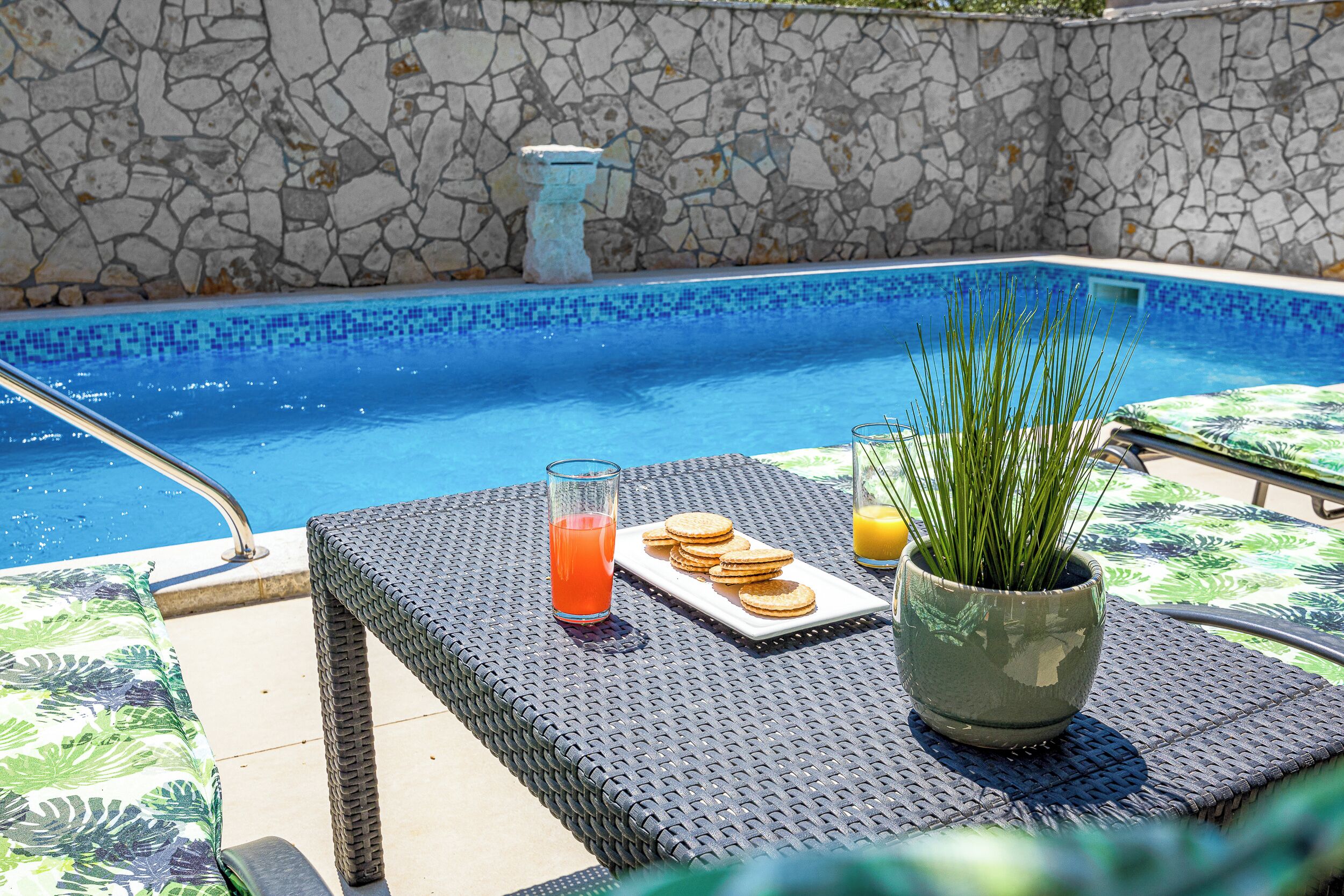 Un espace aménagé au bord de la piscine, avec un mur en pierre, un verre de boisson à l’orange, une assiette de biscuits et une plante en pot.