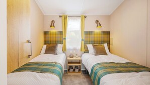 A double room with two beds, a small table, a nightstand, a window with curtains, and two wall-mounted lamps.