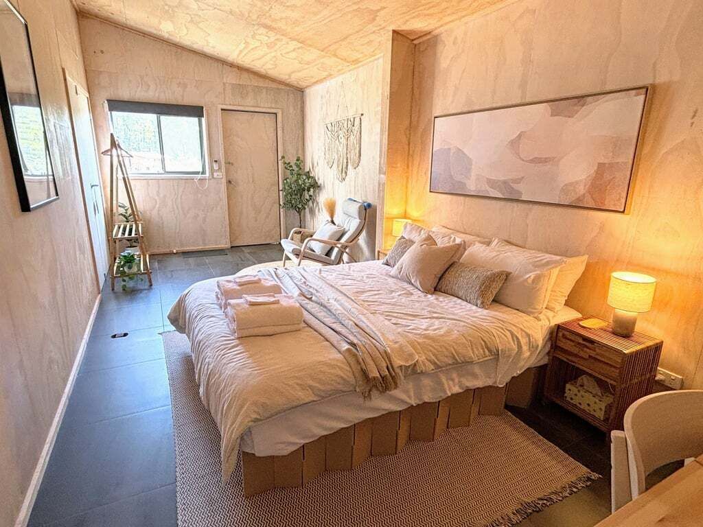 A bedroom with a bed, a chair, a bedside table with a lamp, a wall-mounted artwork, and a door.