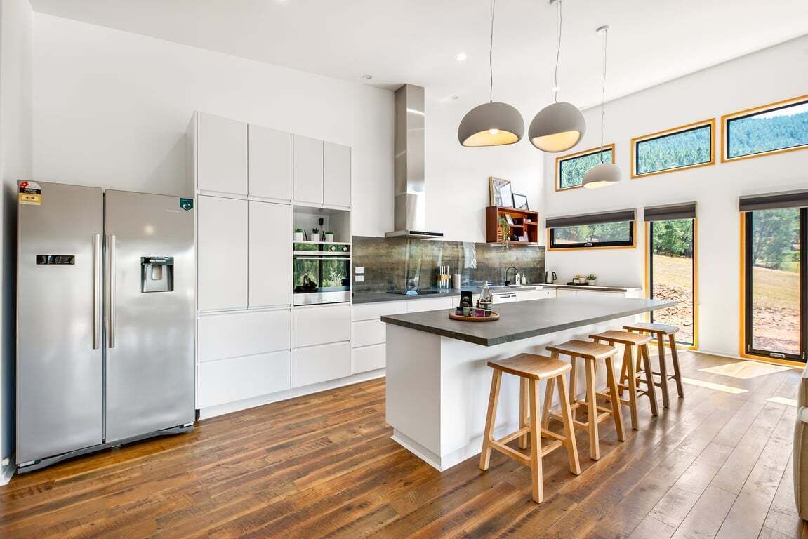 A modern kitchen with white cabinetry, a central island with stools, stainless steel appliances, and large windows.