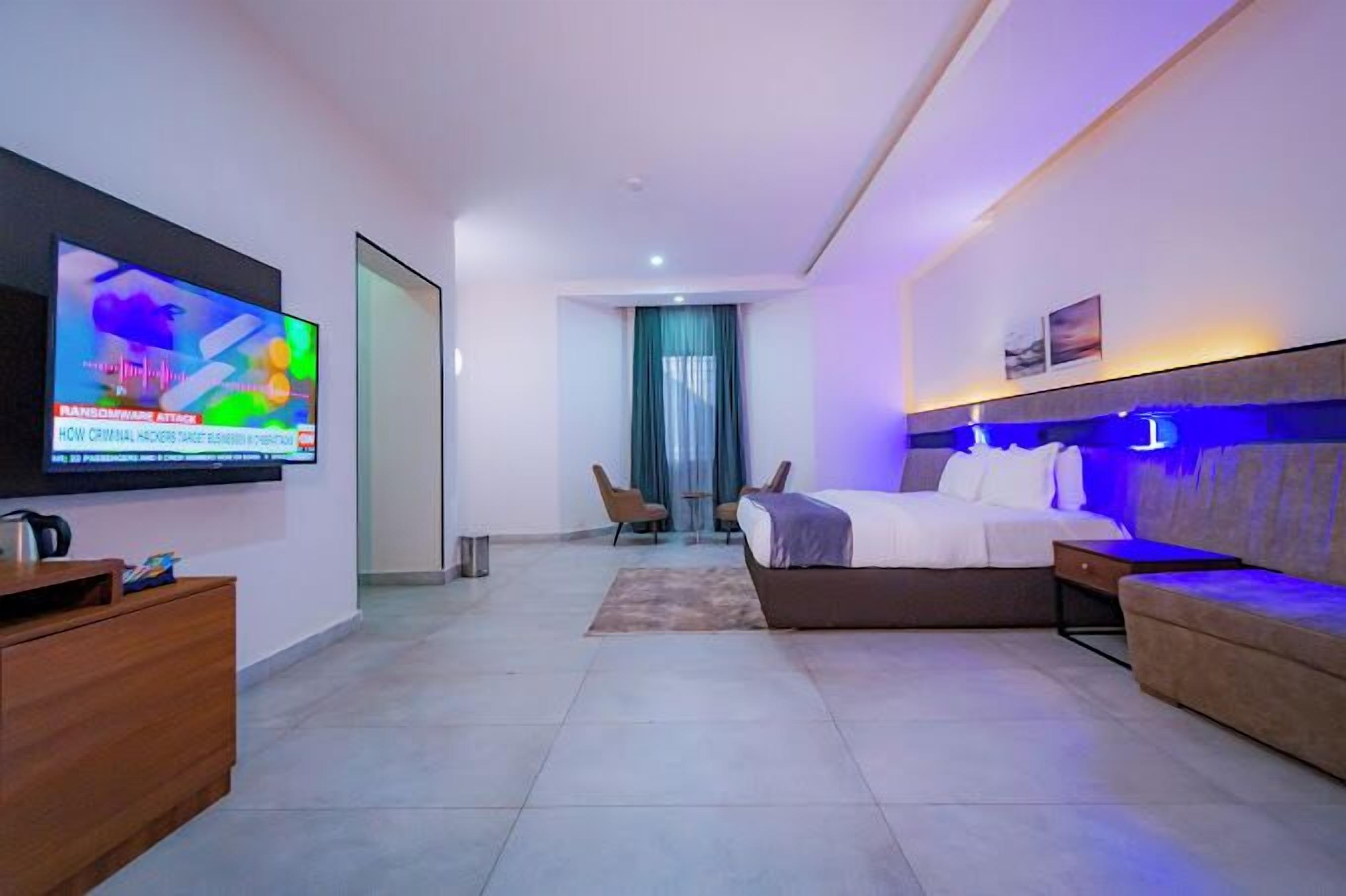 A modern hotel room with a large flat-screen TV, a bed, a sofa, and a desk.