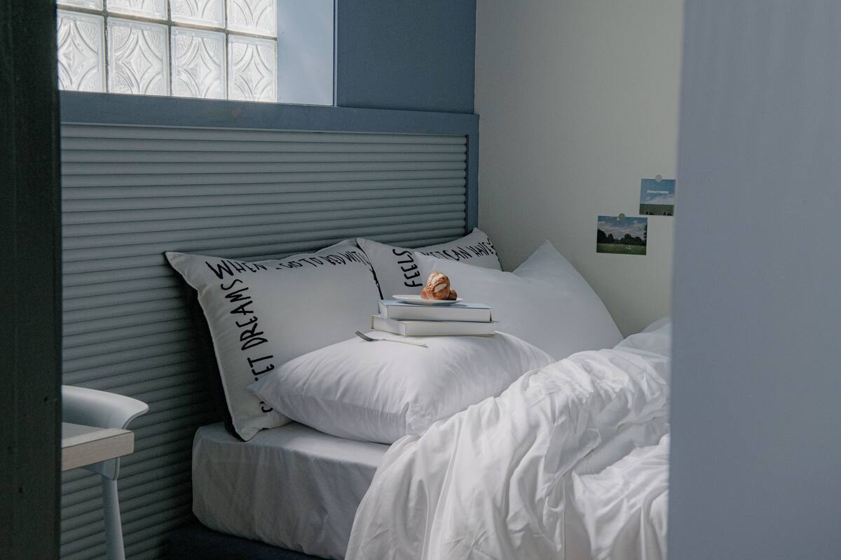 Cama bem arrumada com roupa de cama e almofadas brancas, uma pequena pilha de livros e um objeto decorativo sobre as almofadas.