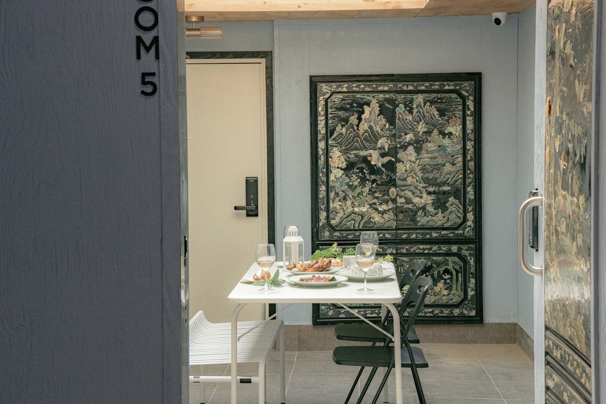 Uma área de refeições com uma mesa posta para dois, uma porta decorativa com arte tradicional e uma placa indicando a sala número 5.