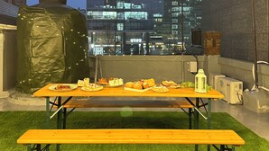 En tagterrasse med et træbord dækket til spisning, omgivet af kunstgræs, med udsigt over en by med moderne bygninger.
