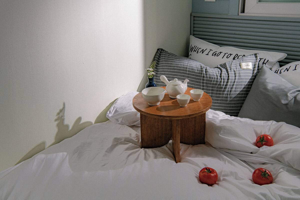Cama com roupa de cama branca, mesa de cabeceira de madeira com conjunto de chá e almofadas com texto.