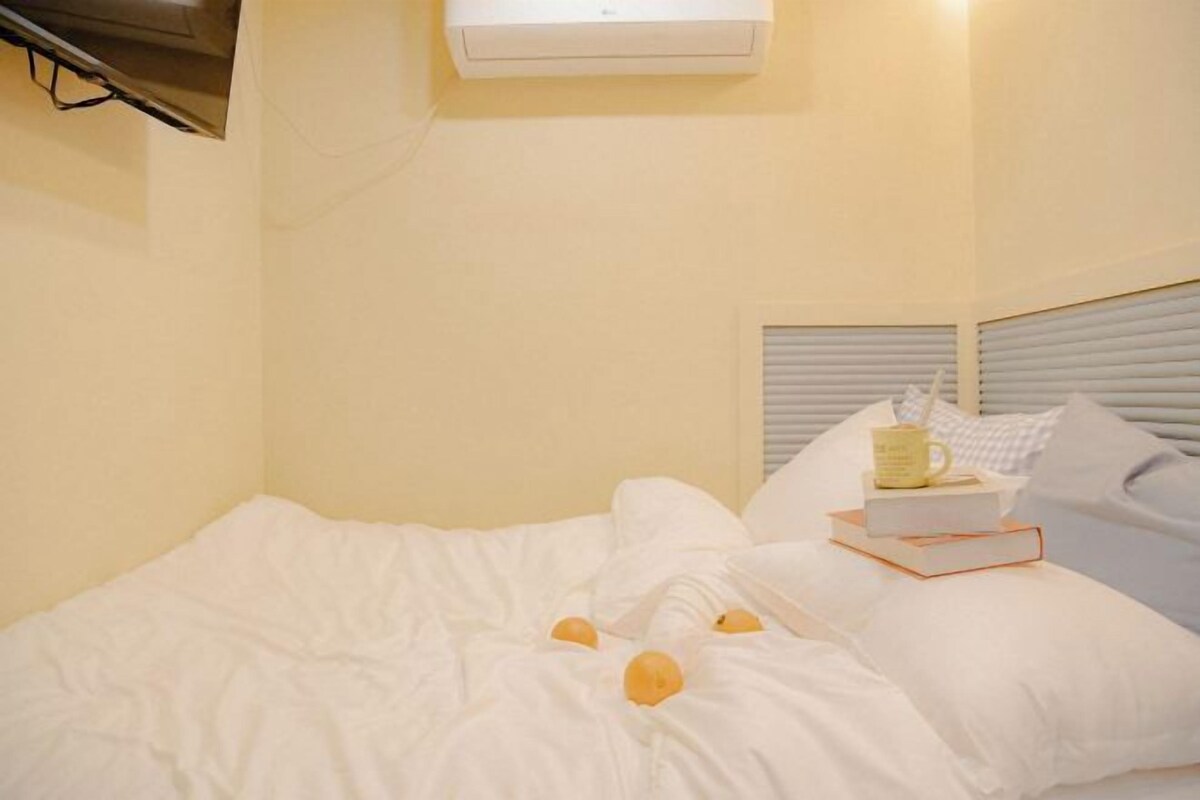 A bedroom with a bed, white bedding, a bedside table with a book and a mug, and a window with blinds.