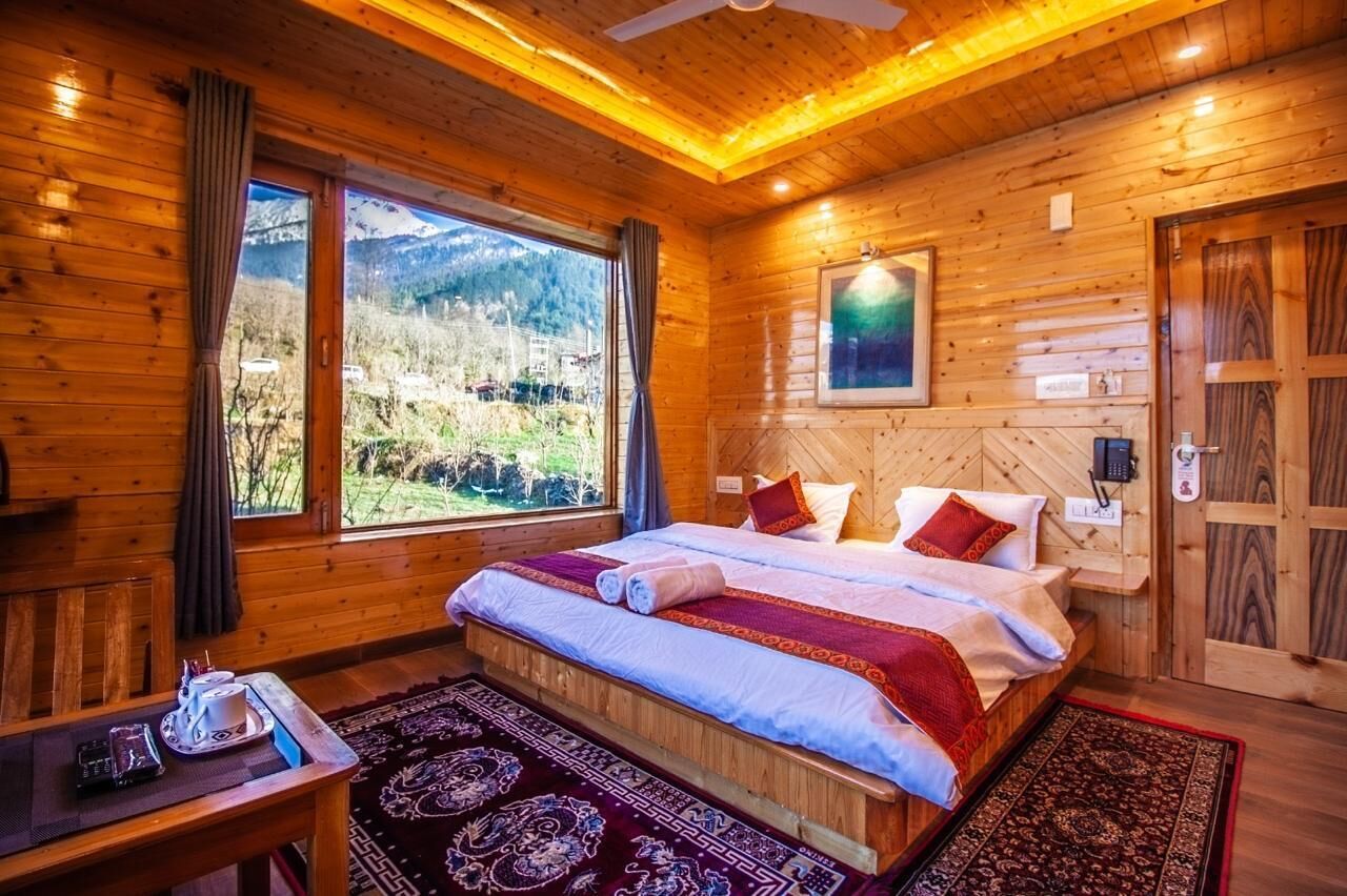 A wooden cabin bedroom with a bed, a table with a cup and saucer, and a view of mountains through large windows.