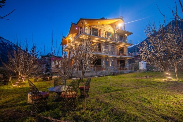 A multi-story building with a stone facade, outdoor seating area with metal chairs, and a well-lit entrance. - Shalai Resort & Cottages (Manali)