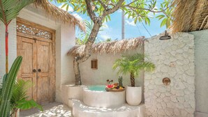 A stone-walled spa area with a wooden door, a thatched roof, and a small pool with fruit.