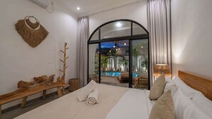 A bedroom with a large bed, a wooden bench, a woven wall decoration, and a view of an outdoor pool area.