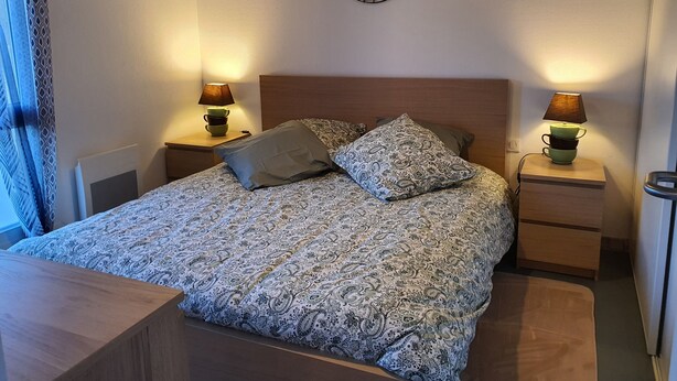 Una cama bien hecha con una colcha estampada, dos lámparas de noche, una obra de arte en la pared y una ventana con cortinas con diseño.