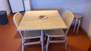 Una mesa cuadrada de madera con dos sillas de mimbre, una mesita redonda y un cubo de basura.