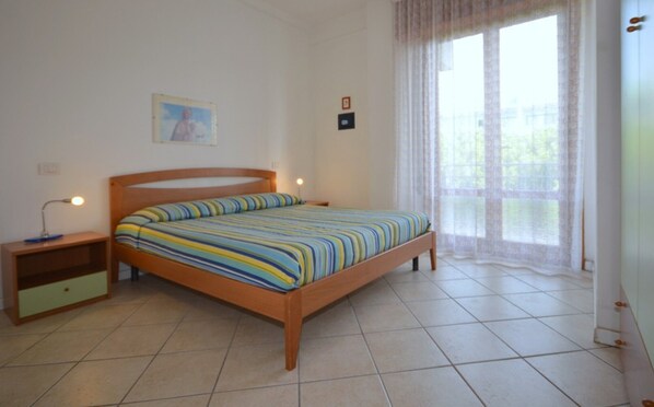 A bedroom with a bed, a nightstand, a lamp, a window with curtains, and a wall-mounted picture. - Spacious apartment 50mt from the beach (Bibione)