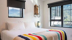 A bedroom with a bed, two windows, a wall-mounted light, and a view of the outdoors. - Breathtaking Condo with Panoramic Mountain Views (Revelstoke)