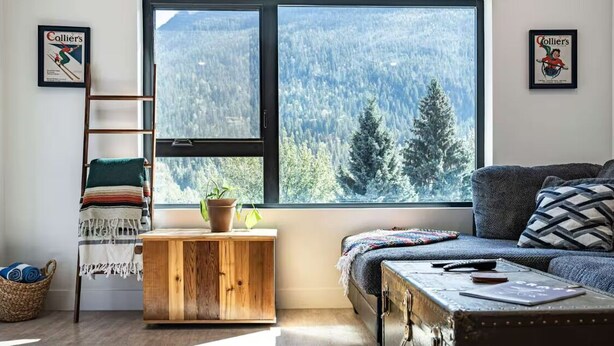 A cozy living room with a large window offering a forest view, a wooden cabinet, a cushioned sofa, and a small table with a plant. - Breathtaking Condo with Panoramic Mountain Views (Revelstoke)