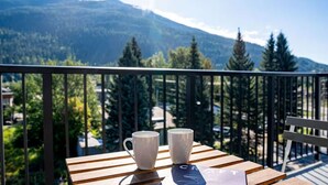 A balcony with two white mugs on a wooden table, overlooking a forested mountain area. - Breathtaking Condo with Panoramic Mountain Views (Revelstoke)