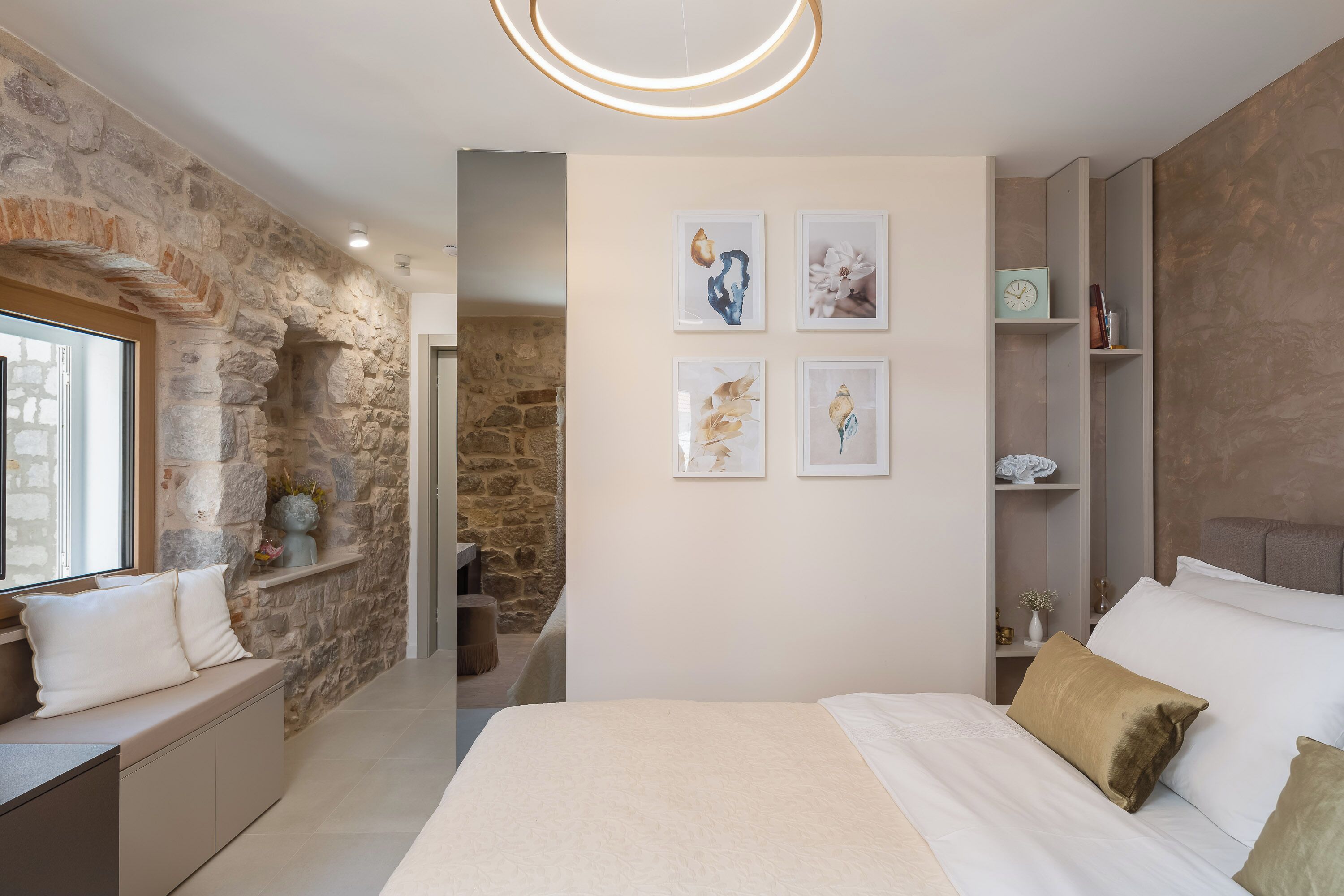 Une chambre avec un mur en pierre, un lit avec une literie blanche et beige, et quatre œuvres d’art encadrées accrochées au mur.