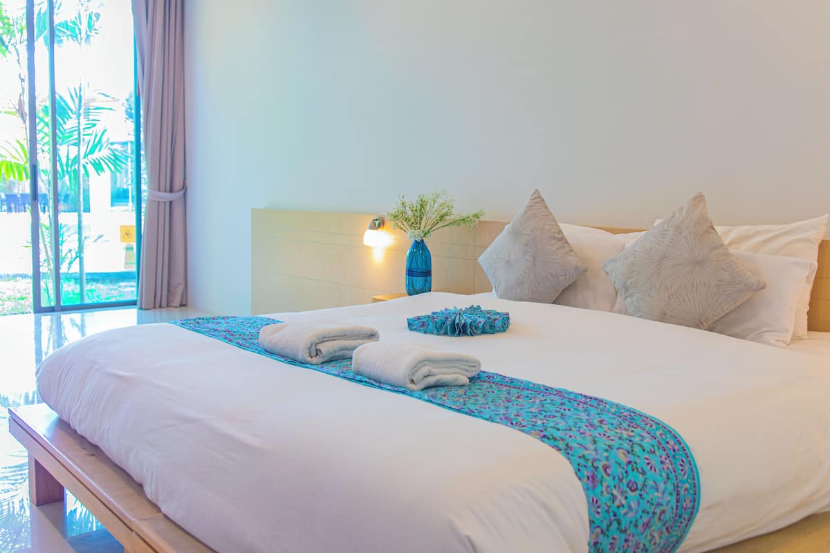 A neatly made bed with a blue patterned runner, two pillows, and a vase with flowers on a nightstand.