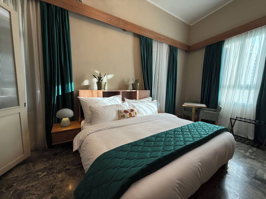 A hotel room with a large bed, a bedside table with a lamp, a small table with a chair, and a window with curtains.