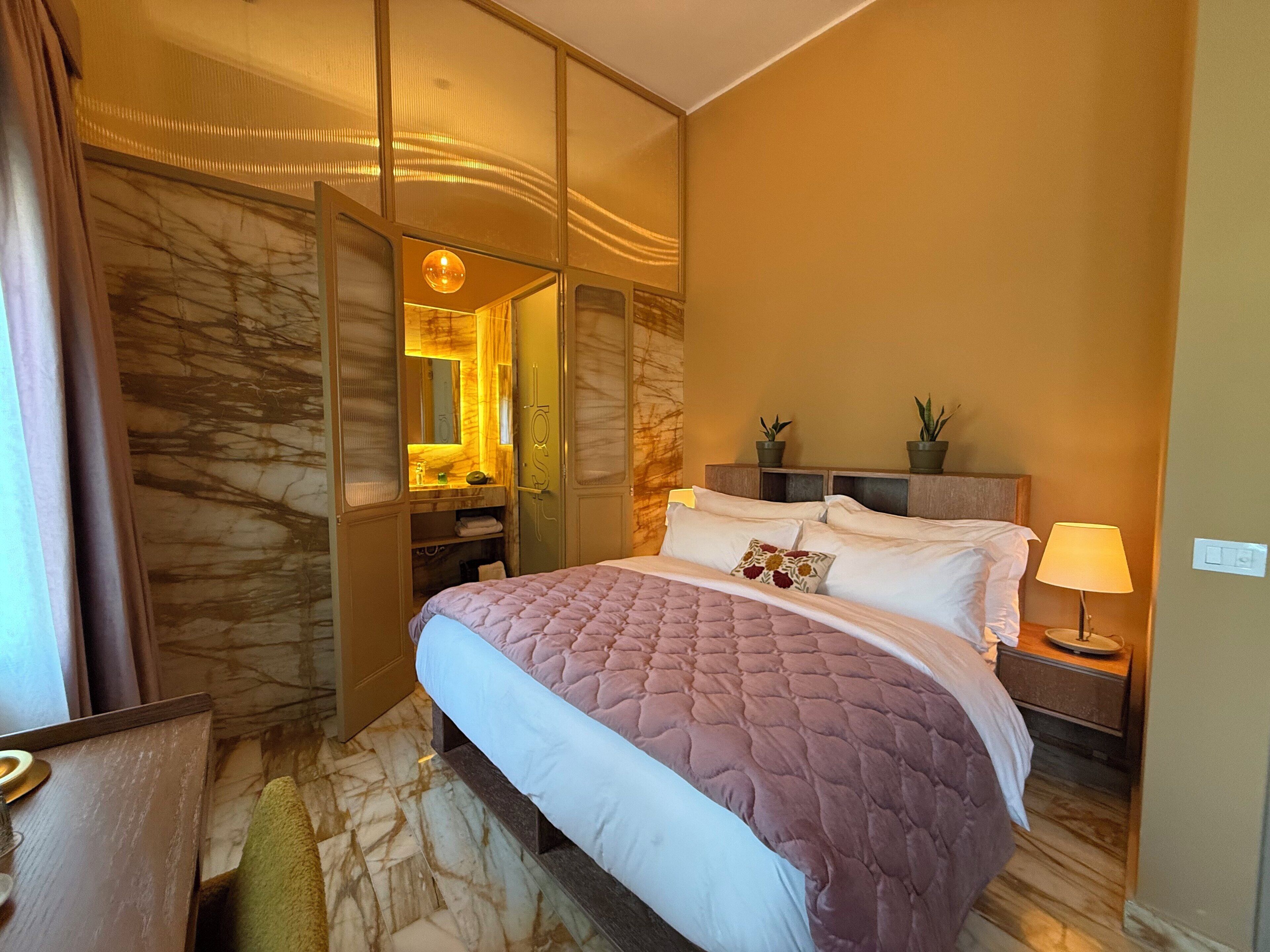 Habitación de hotel con cama, mesita de noche, lámpara, espejo y baño con lavabo y toallero.