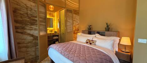 Habitación de hotel con cama, mesita de noche, lámpara, espejo y baño con lavabo y toallero.