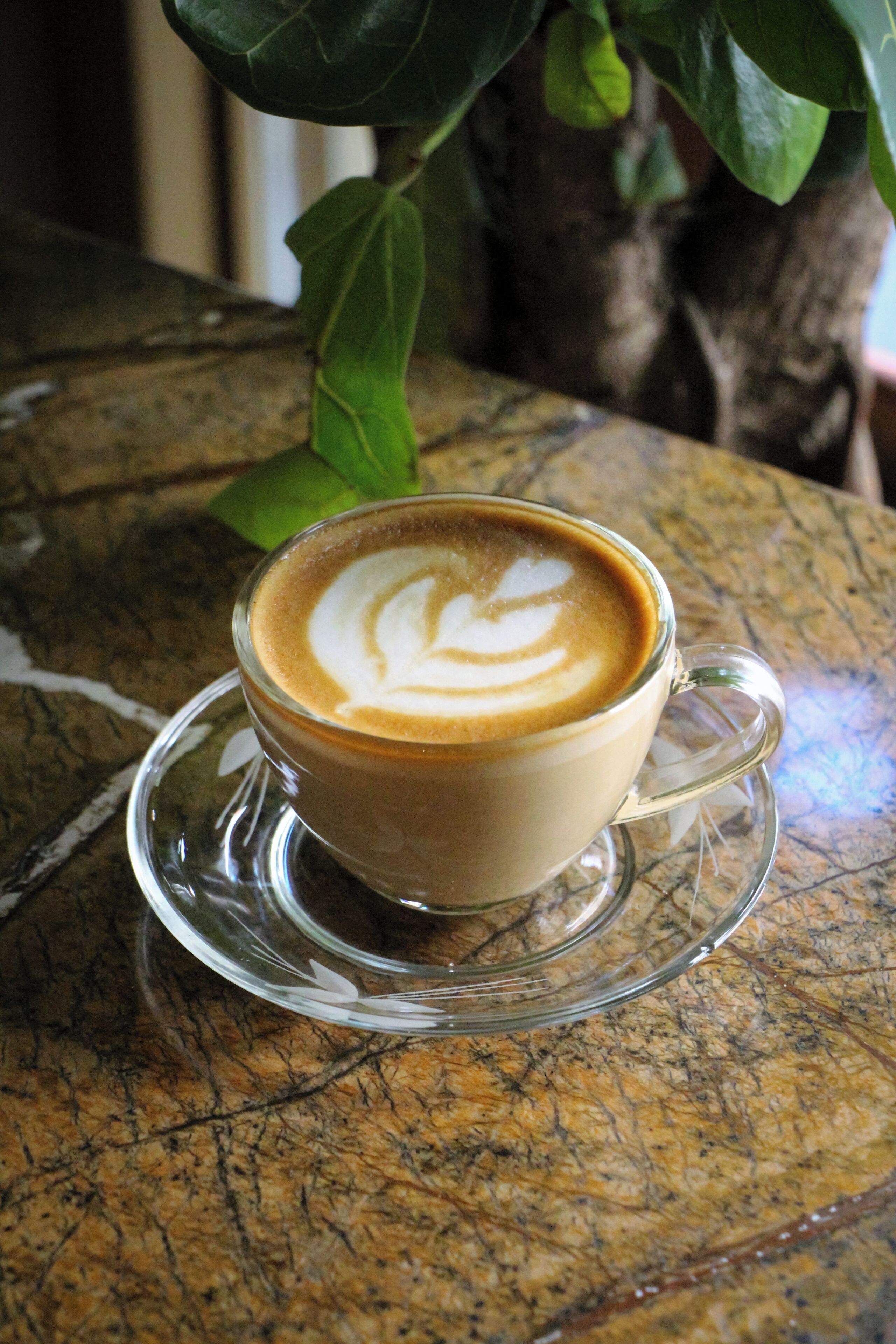 A cup of coffee with latte art on a clear glass saucer on a textured surface.