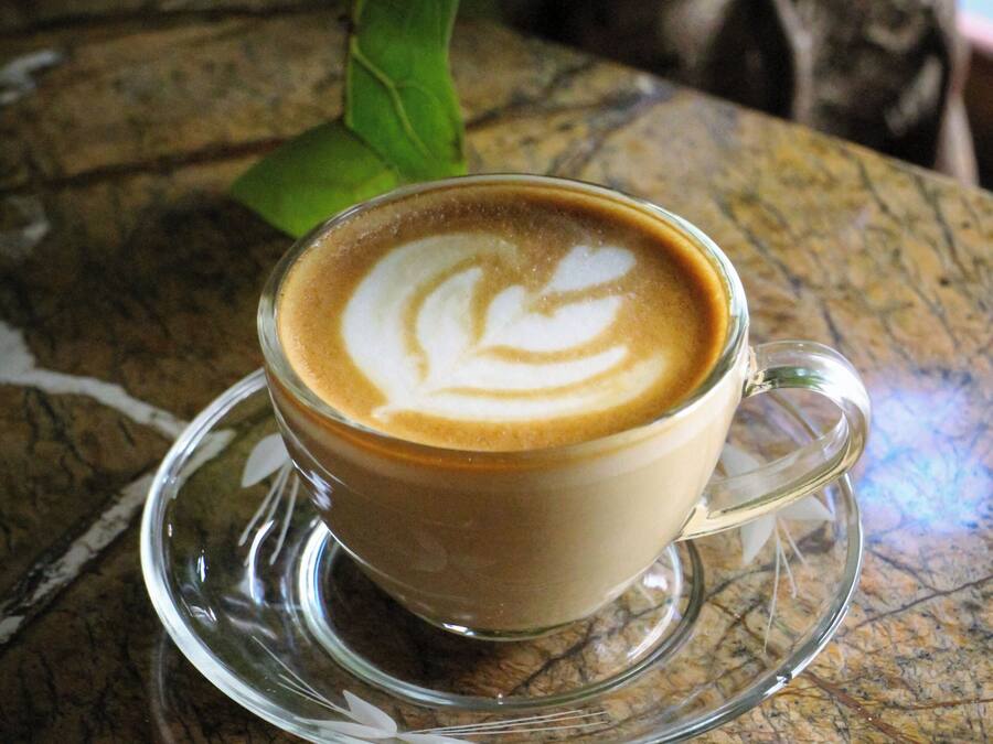 Una taza de café con arte de latte sobre un platillo de vidrio transparente sobre superficie texturizada.