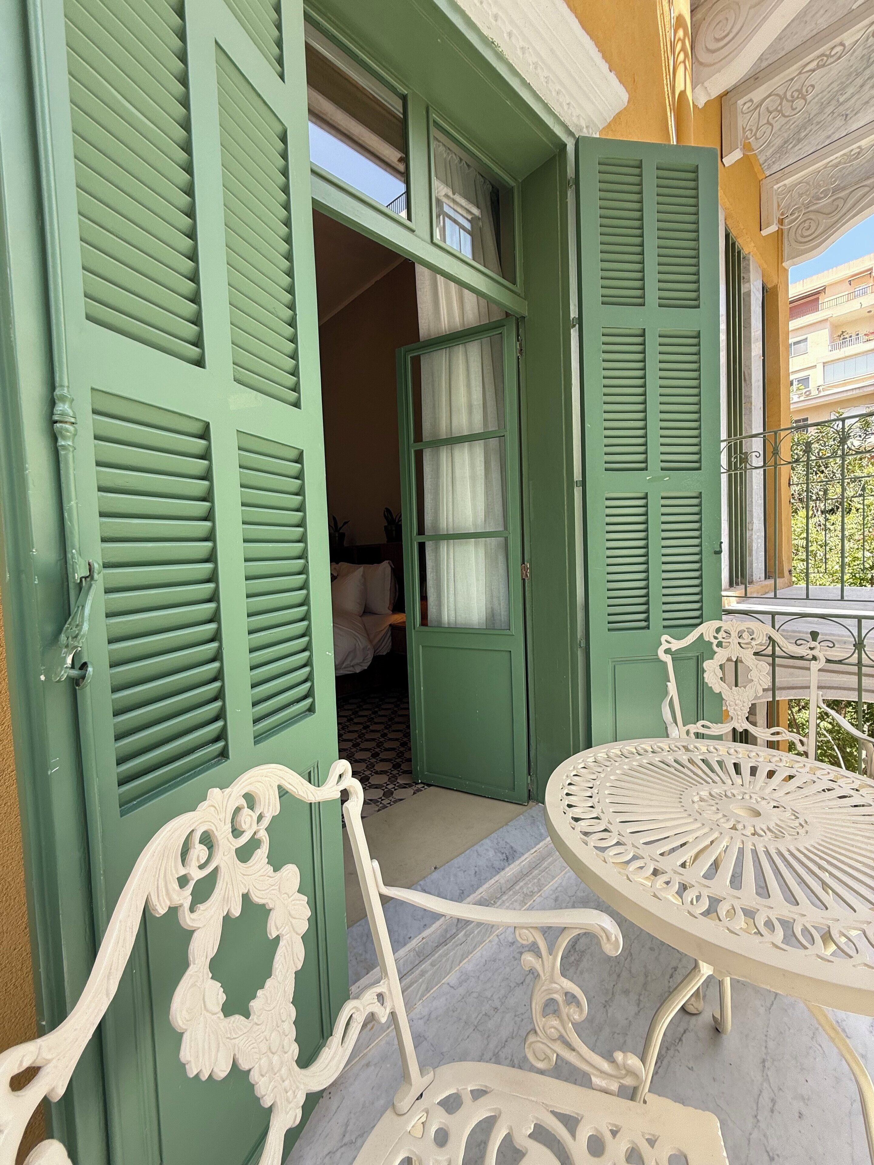 A green door with shutters, an open door revealing a room with a bed and curtains, and a white outdoor table with chairs.
