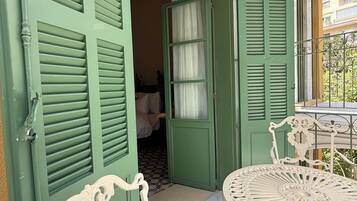Una puerta verde con contraventanas, una puerta abierta que muestra un cuarto con cama y cortinas, y una mesa blanca al aire libre con sillas.