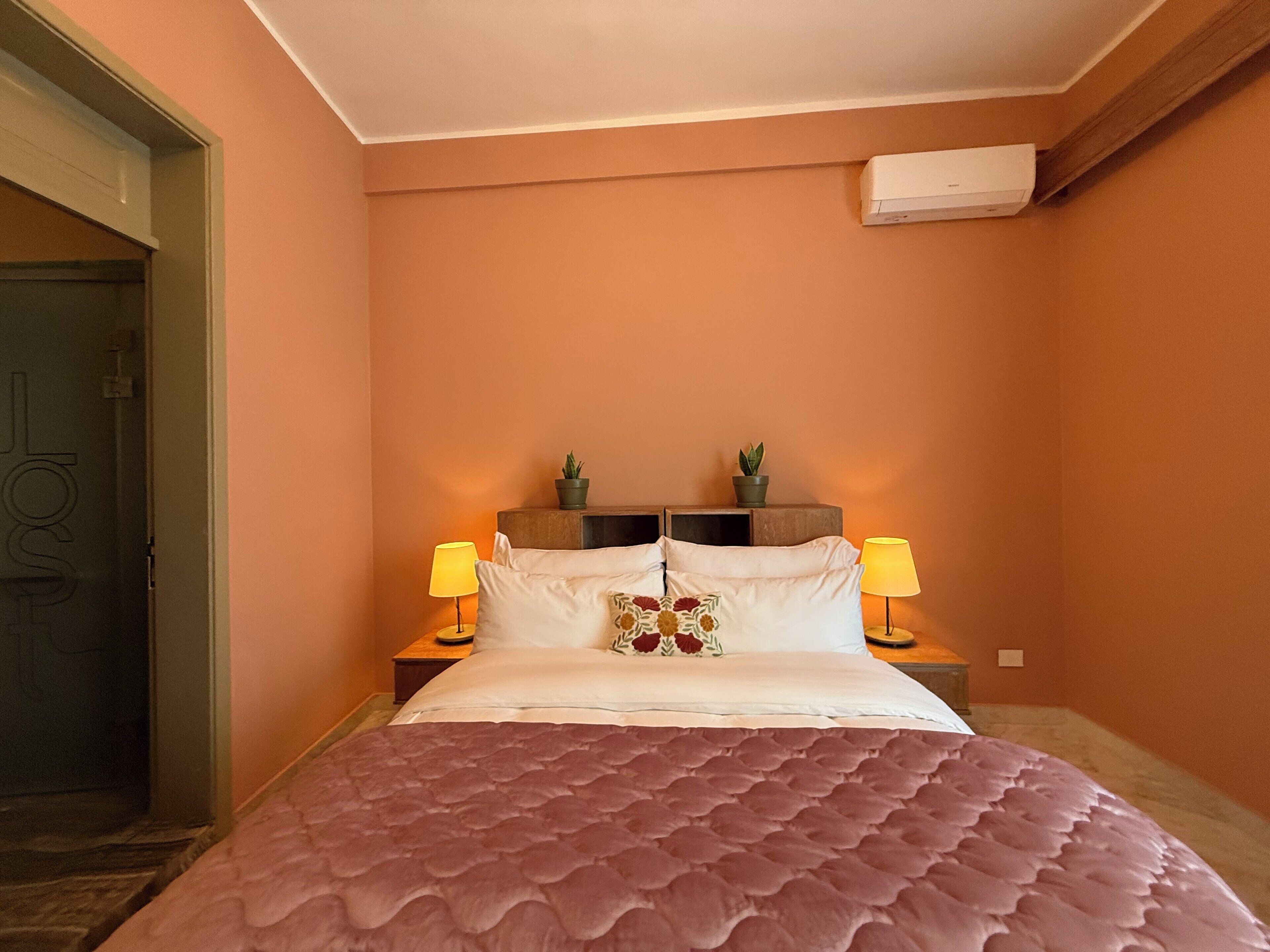 Un dormitorio con una cama grande, dos mesitas de noche con lámparas, una planta en maceta sobre cada una y un equipo de aire acondicionado en la pared.