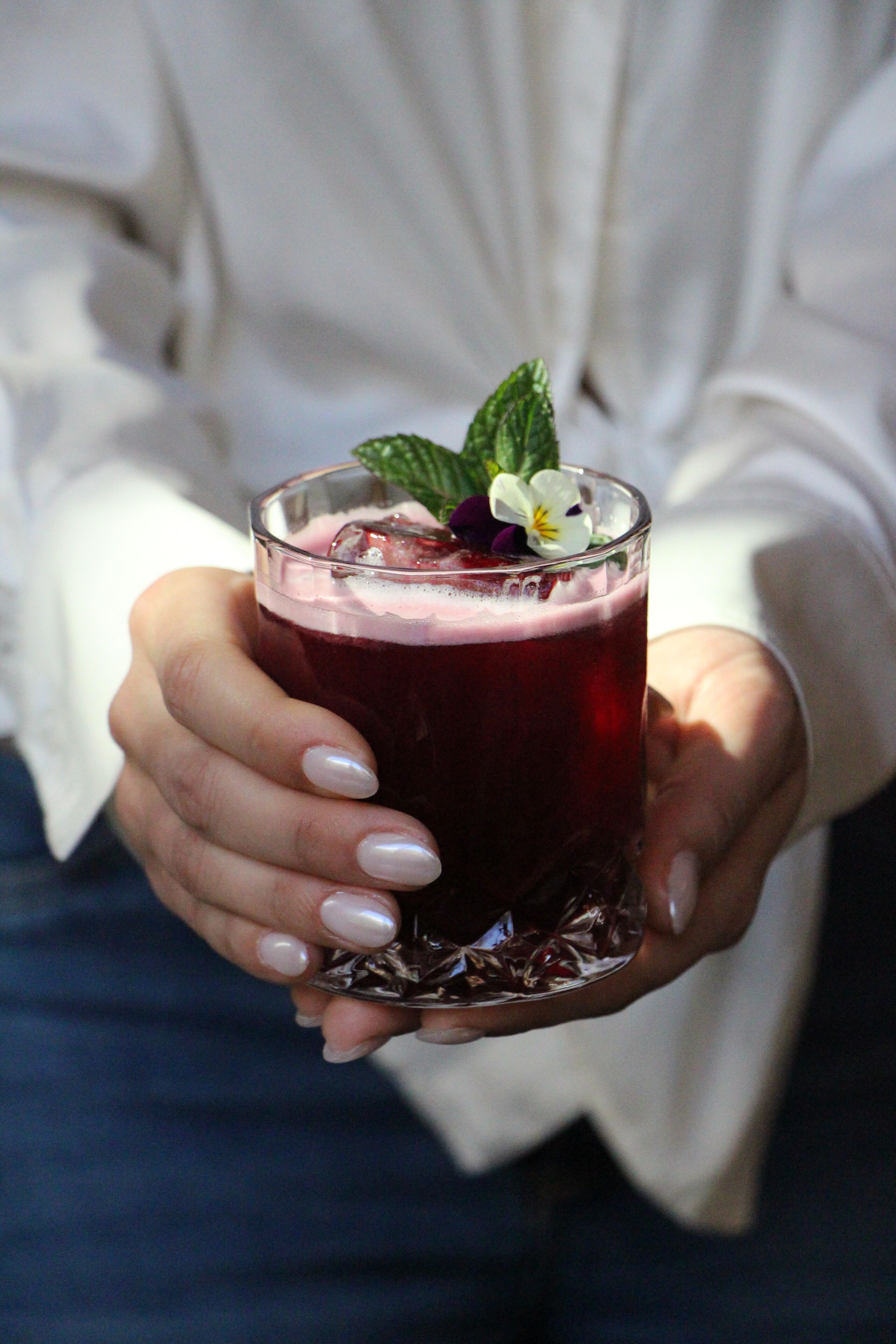A person holding a glass of red beverage with ice cubes and a mint garnish.