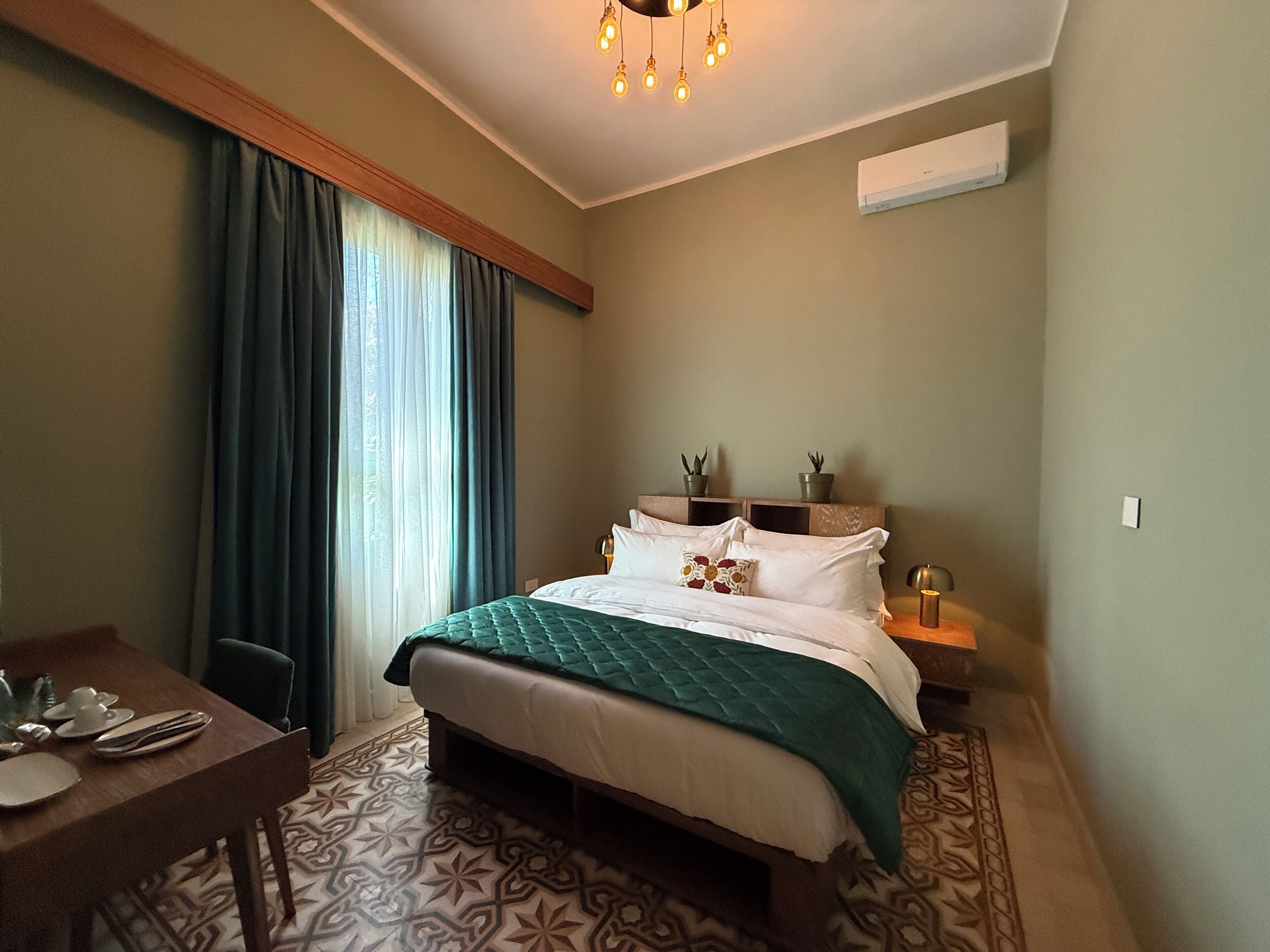 A hotel room with a bed, a desk, and a chair. There is a window with curtains, a ceiling light, and an air conditioning unit on the wall.