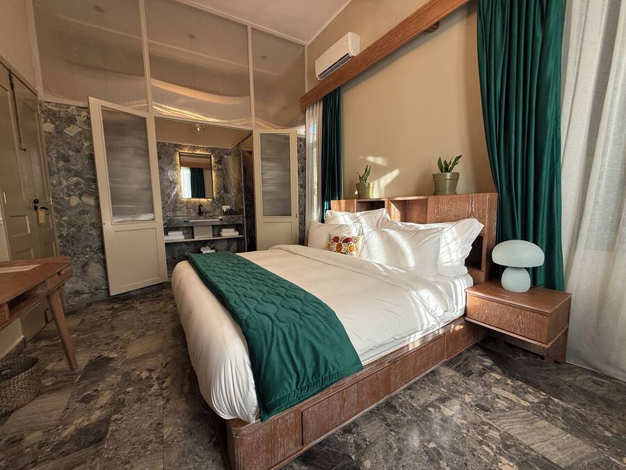 Een hotelkamer met een groot bed, een nachtkastje van hout, een spiegel, een badkamer met een marmeren wand en een groen gordijn.