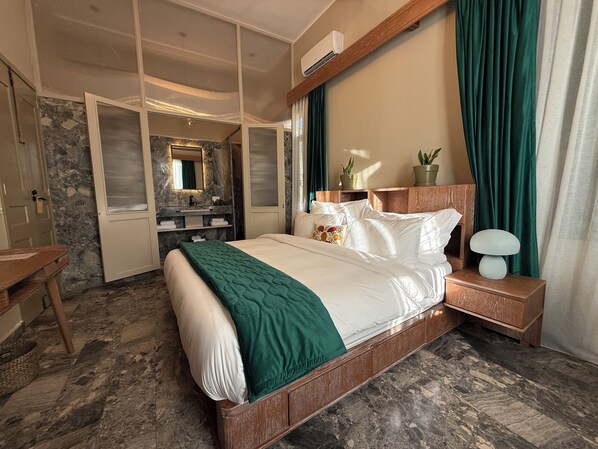 A hotel room with a large bed, a wooden nightstand, a mirror, a bathroom with a marble wall, and a green curtain. - Hotel Lost Ashrafieh (Beirut)