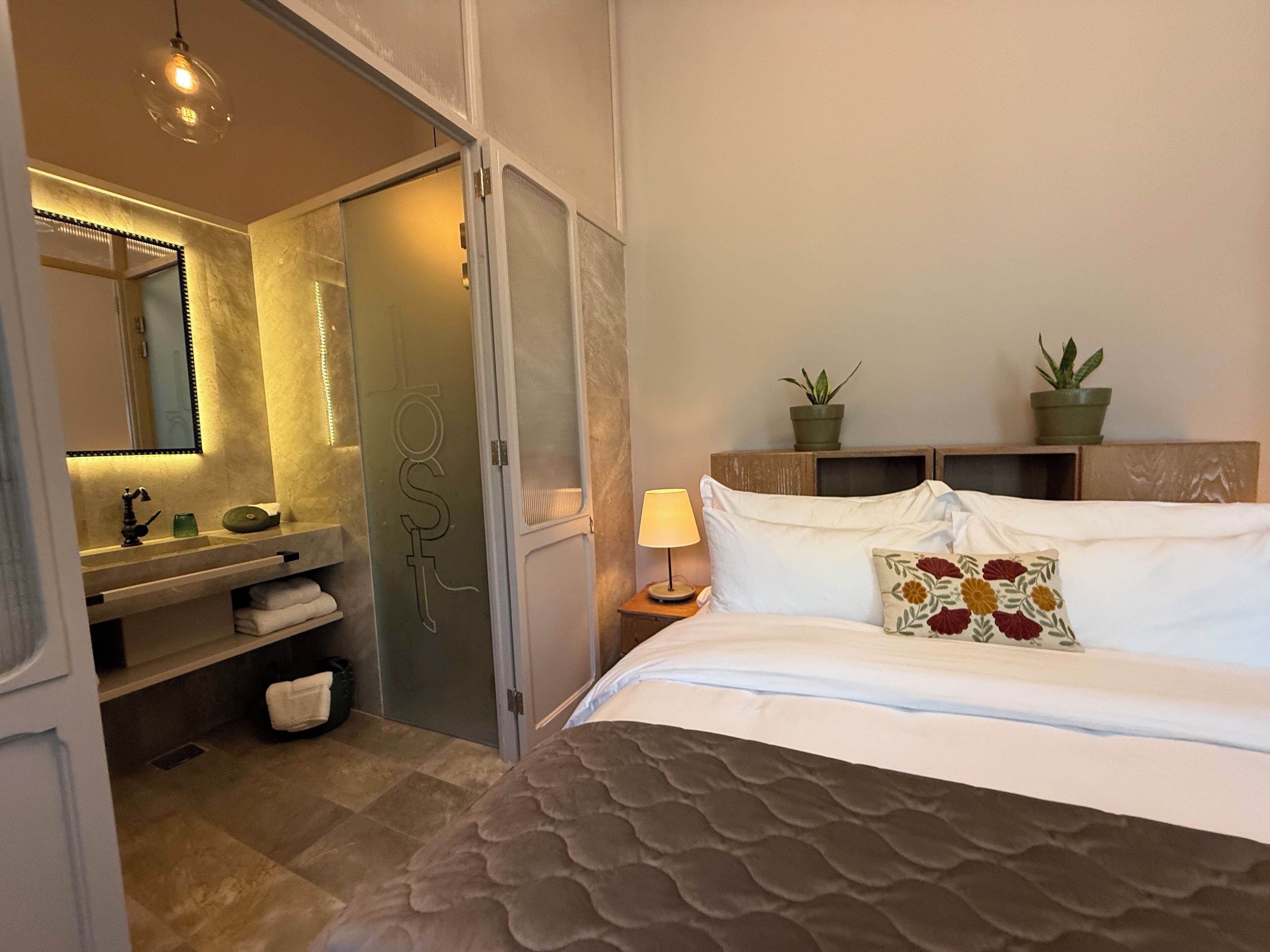 A hotel room with a bed, a nightstand, a lamp, a mirror, a bathroom with a sink, and two potted plants.