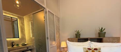 A hotel room with a bed, a nightstand, a lamp, a mirror, a bathroom with a sink, and two potted plants.