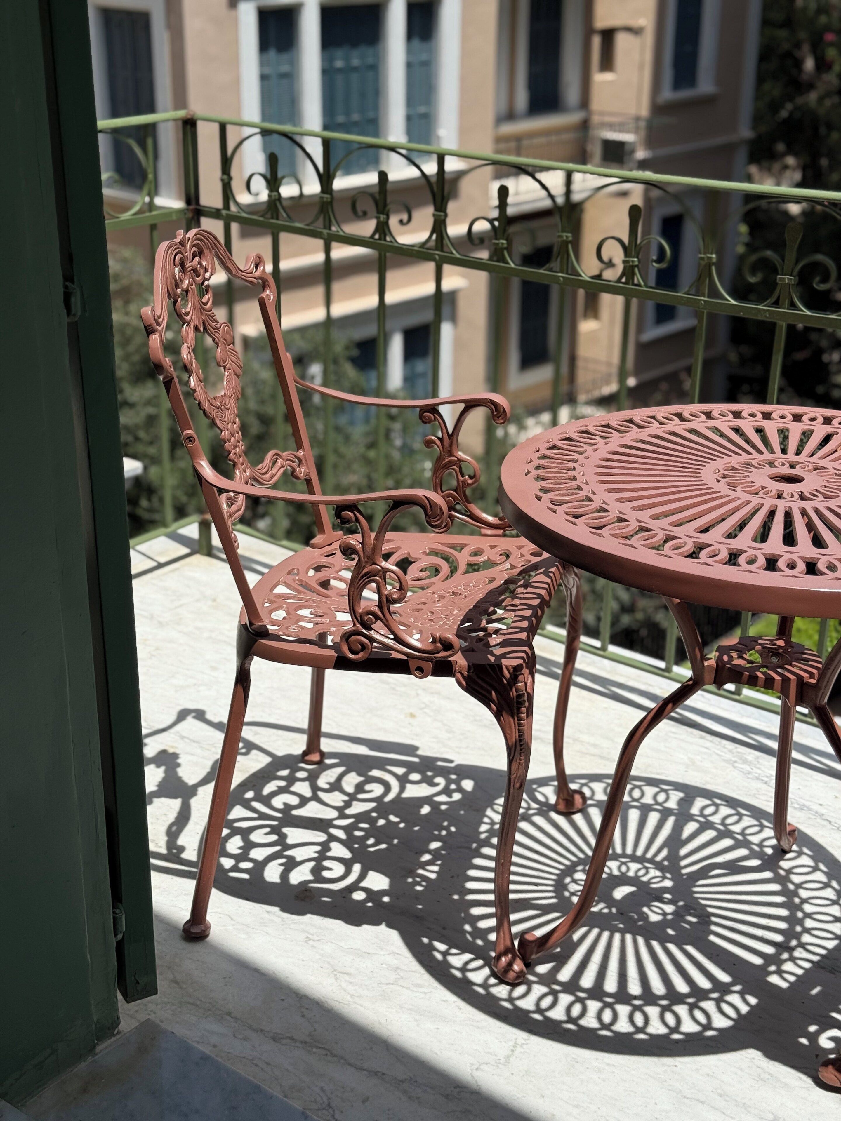 A wrought iron balcony table and chair set with intricate designs, positioned on a balcony with a view of buildings in the background.