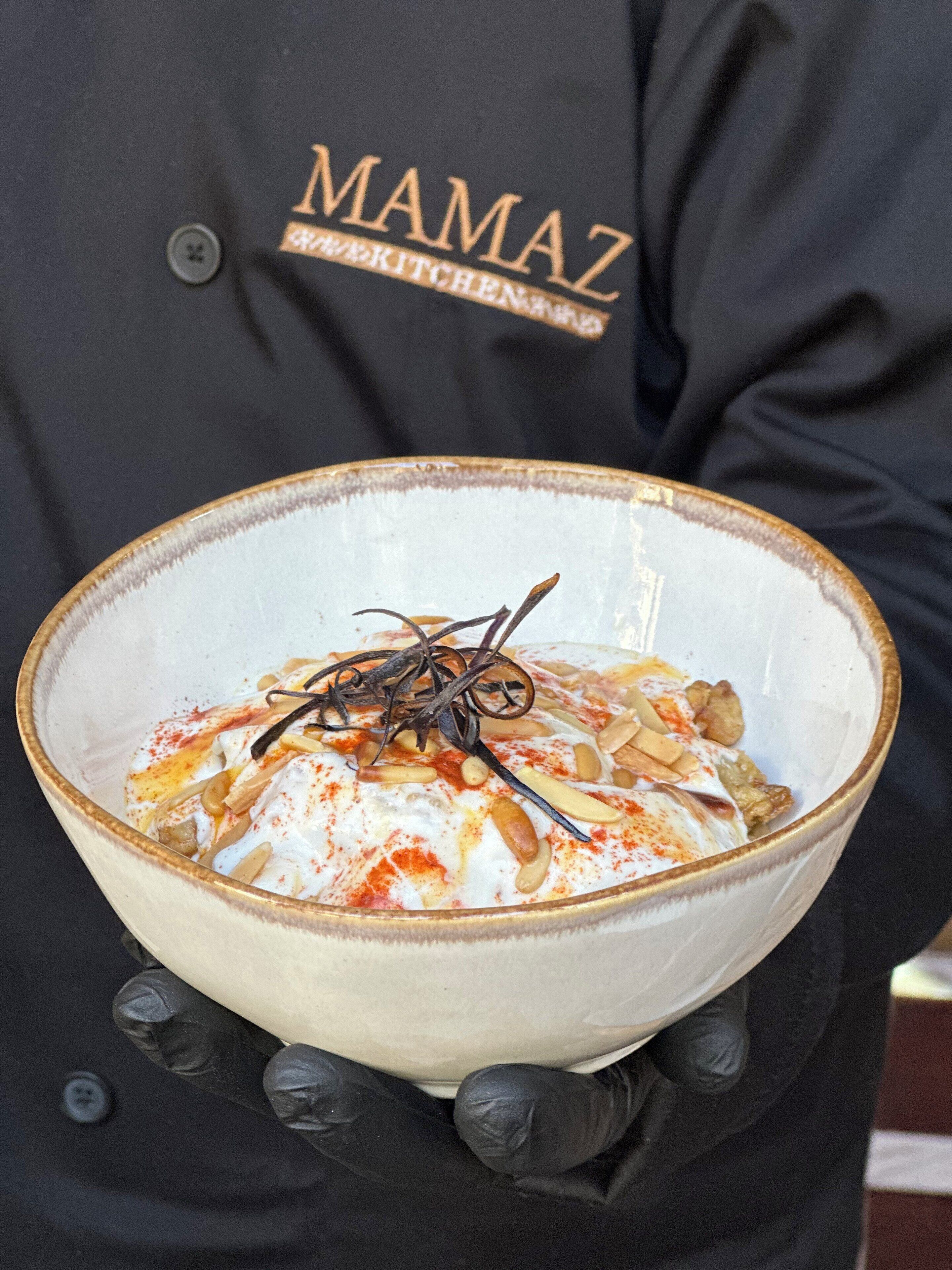 Una persona sosteniendo un tazón de comida con diferentes ingredientes, usando un delantal negro con la inscripción 'MAMAZ'.