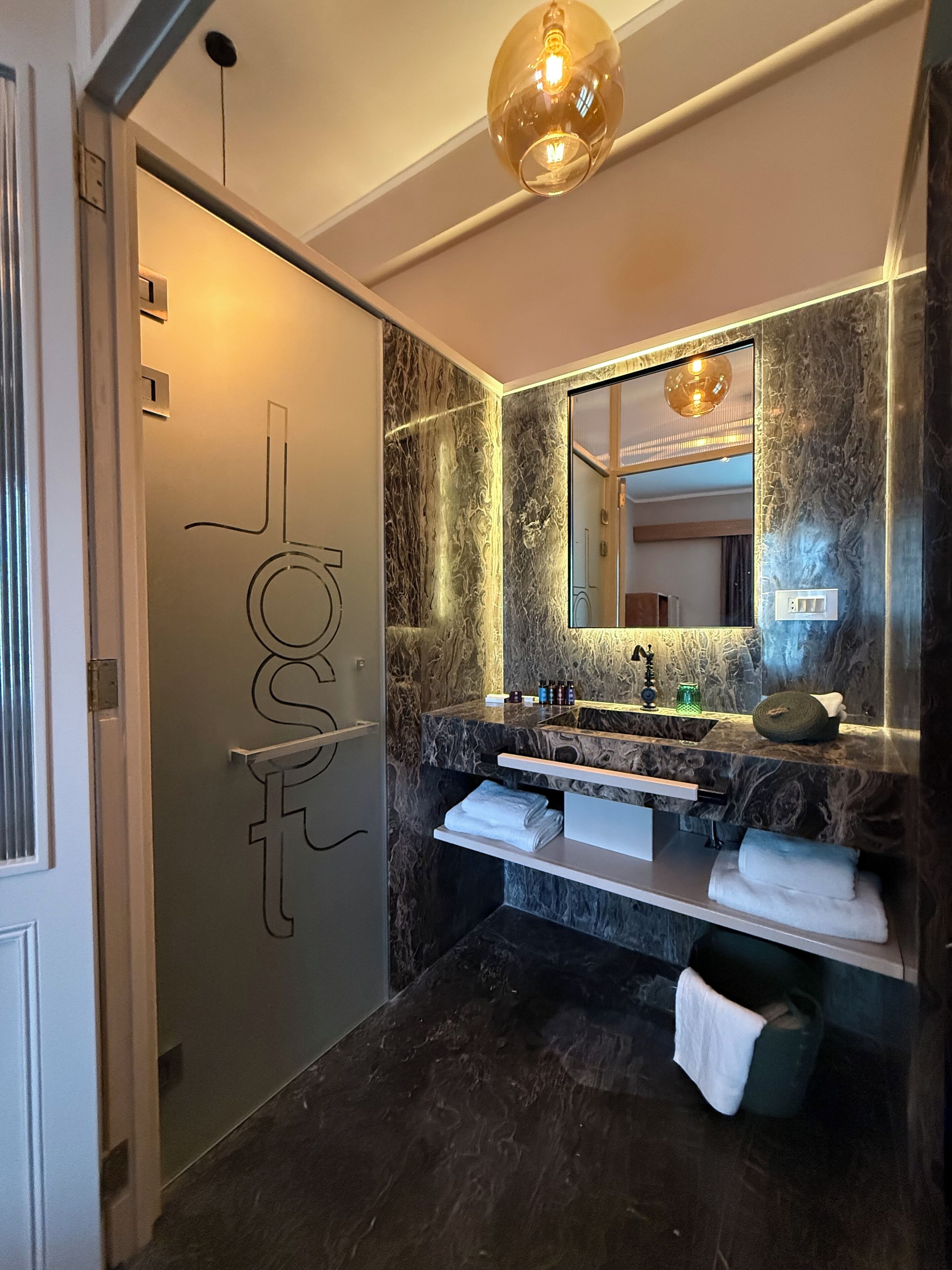 Un baño moderno con tocador de mármol, un espejo grande y una puerta de vidrio con una lámpara colgante.