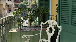 Un balcon avec une chaise et une table en métal peintes en blanc, des volets verts et une vue sur des immeubles et des arbres.