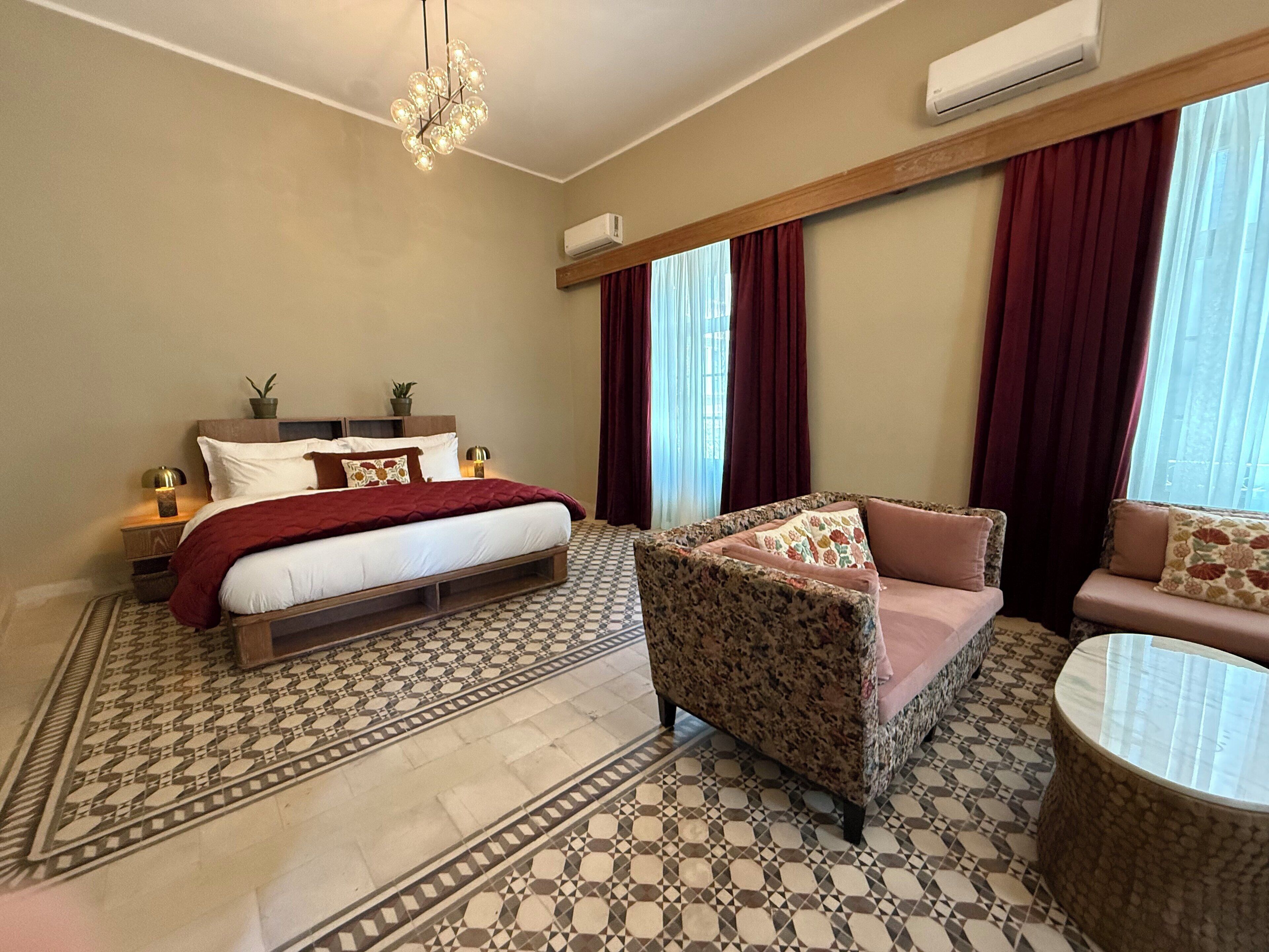 A bedroom with a bed, a sofa, a chair, a patterned floor, and a chandelier.