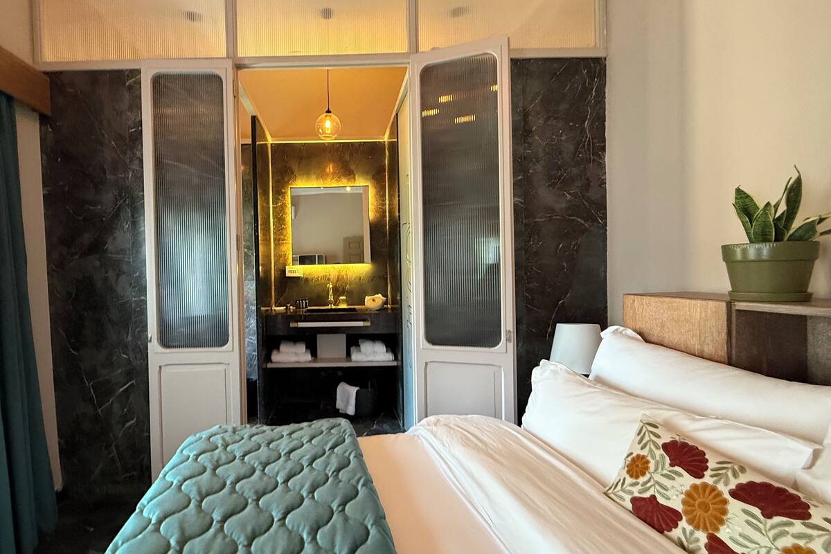 Une chambre d’hôtel avec un lit, une table de chevet ornée d’une plante, une salle de bain avec un miroir et un rideau.