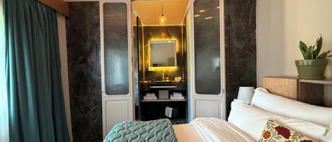 A hotel room with a bed, a bedside table with a plant, a bathroom with a mirror, and a curtain.