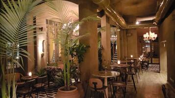 An indoor dining area with tables and chairs, surrounded by large potted plants and a patterned floor.